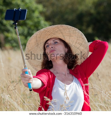 mature selfie concept - sexy aging woman enjoying taking a selfie with a stick in summer dry meadows wearing a summer hat,natural daylight