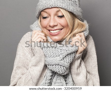 trendy warm winter - gorgeous young blond woman wrapping up herself in gray wool winter hat and scarf smiling for softness and cozy fashion