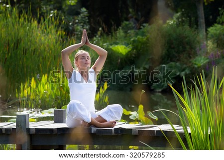 outdoors meditation - relaxed young yoga woman in praying lotus pose,closing eyes to relax and meditate on a wooden bridge with green foreground and water background, summer daylight