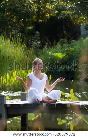 outdoors relaxation - focused young yoga woman in lotus pose,breathing and meditating on a wooden path with green foreground and water lily background, summer exotic daylight