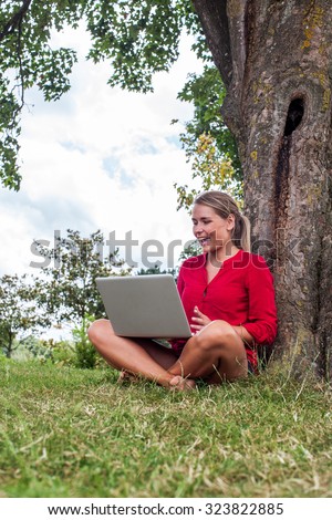 working outdoors - beautiful suntanned 20s girl laughing with laptop on crossed legs sitting under tree working as freelancer,natural summer daylight