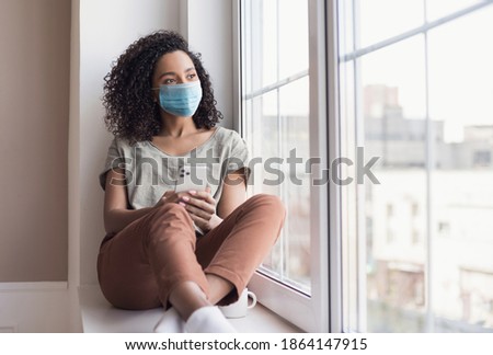 Sad woman alone during coronavirus pandemic wearing face mask indoors at home for social distancing. Mixed race girl looking at window. Anxiety, stress, lockdown, mental health crisis concept