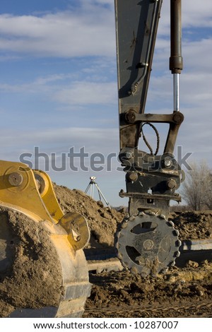 Road construction - excavator arm with a roller to compact the fill over newly placed sewer pipe, backhoe scoop, survey equipment