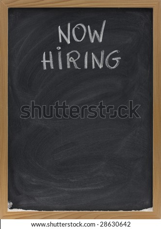 now hiring advertisement handwritten with white chalk on blackboard, copy space below, eraser smudges