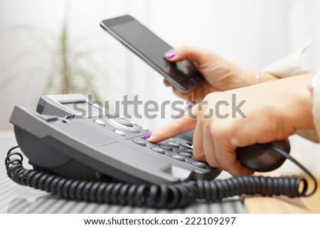 Woman is dialing on land line phone and looking on mobile phone