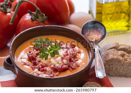 Tomato salmorejo soup in a ceramic bowl with the raw ingredients, tomatoes, eggs, oil and bread over white background