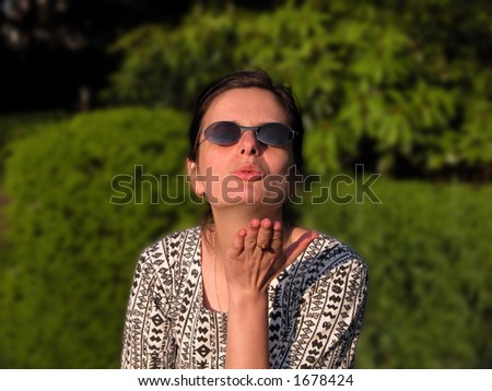 Girl with sunglasses blowing a kiss to you. Selective focus on the face, blurred  background, natural lighting from left with shadows on right.