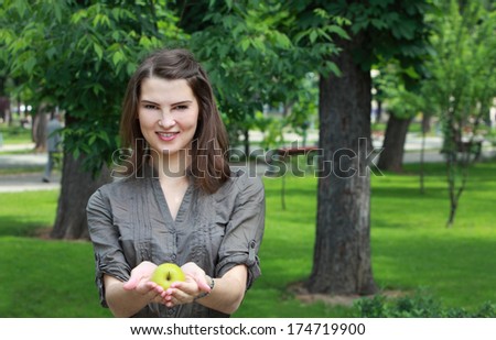 Young smiling woman offering a green apple outside in a park in summer.