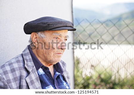 Serious senior man in checkered jacket and cap sitting at fence while looking sideways