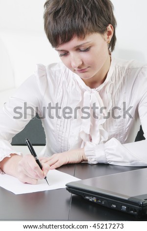 Pretty young businesswoman sitting at desk and writing on blank paper