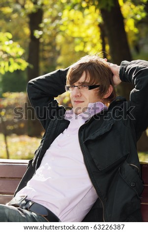 Young pensive man wearing casual clothes sitting in park on bench
