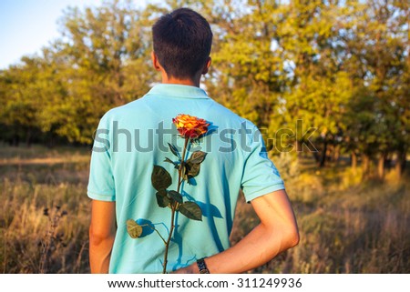 man holding a rose behind his back in a park outdoors