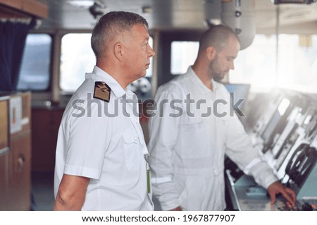 The captain of a cargo ship in a white shirt and shoulder straps on the bridge gives instructions to the navigator Foto stock © 