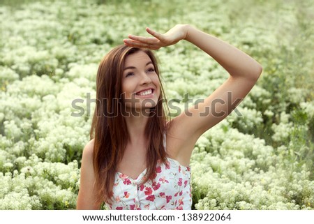 Portrait of beautiful smiling female model at the field of white flowers, looking up with hand over her forehead