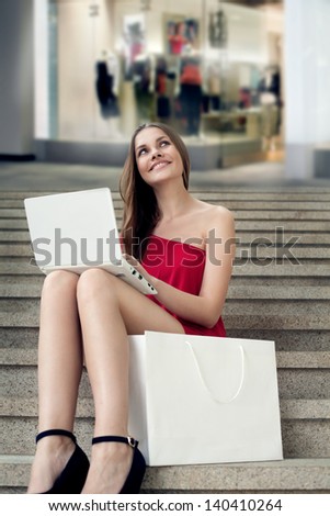 Beautiful woman is surprised looking at her laptop, while sitting on the stairs with shopping bag