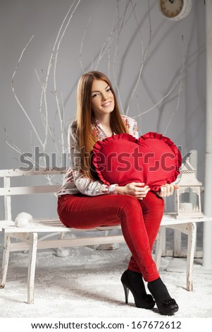 Young woman holding a red heart while sitting on a bench in winter studio interior