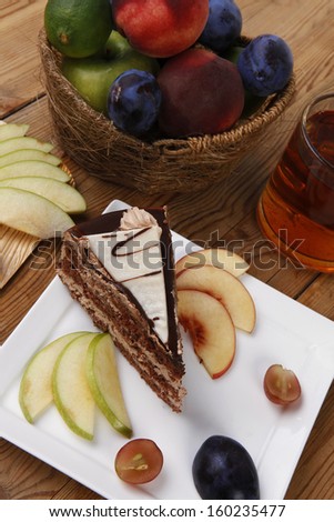 slice of chocolate cream brownie cake topped with white chocolate and cream flowers with hot tea mug decorated with fruits apple plum and grape on plate on wooden table