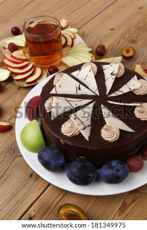 big whole frost chocolate cream brownie cake topped with white chocolate and cream flowers with hot tea cup decorated with fruits apple plum and grape on plate on wooden table