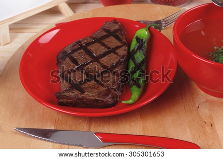 red theme lunch : fresh grilled bbq roast beef steak on red plate with green chili tomato soup ketchup sauce and modern cutlery served on wooden plate over table