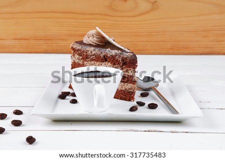 breakfast hot coffee mug and cream chocolate layer cake decorated with white chocolate slice and cream flower on white plate over wood