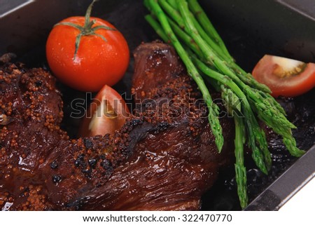 fresh glazed baked big beef meat rib chunk under sweet sauce with tomatoes hot chili pepper pink peppercorn and asparagus in black tray isolated on white background