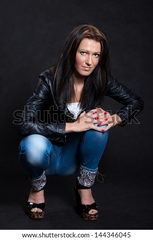 Woman with long hair in black leather jacket and jeans is squatting on a black background