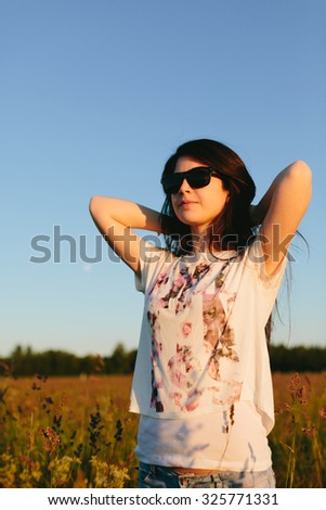 Cute brunette girl in sunglasses in a field. Hands behind her head in a field.