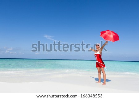 Santa woman standing at the beautiful white sand beach and holding the Christmas red umbrella. Copy space