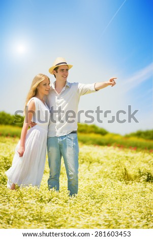 Happy, young couple standing in a chamomile, daisy field.Young man pointing finger at something, looking away