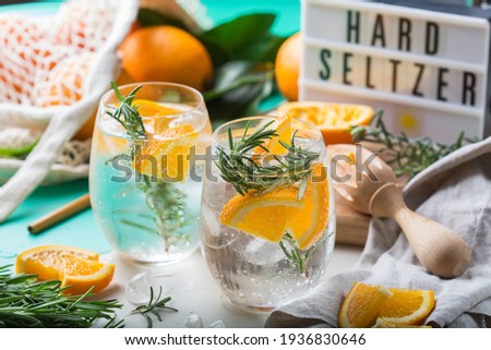 Hard seltzer cocktail with orange, rosemary and ice on a table. Summer refreshing beverage, drink with trendy zero waste accessories, bamboo straw and mesh bag.