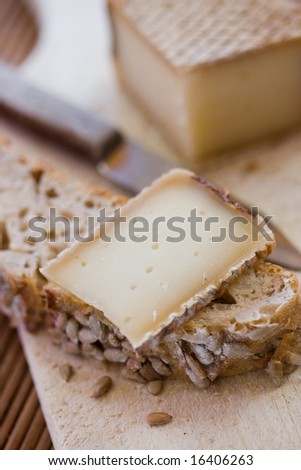A french tasty slice of cheese on a piece of bread with cereals on a wooden board, with the whole cheese and a knife in the background