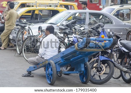 MUMBAI, INDIA - FEBRUARY 10, 2014: Worker sitting on his push cart after delivering goods with busy traffic at background