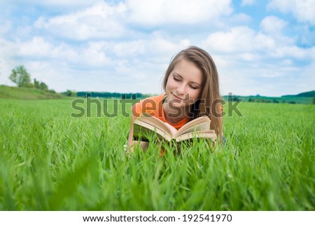 Young nice attentive woman lies on green grass and reads book in a field.