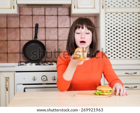 Beautiful funny young woman eating french bread pizza and burger in the kitchen. Happy female eating fast food.