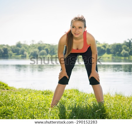 Runner girl outdoors resting on the bank of the lake or river. Tired but happy beautiful sports fitness model living healthy active lifestyle.