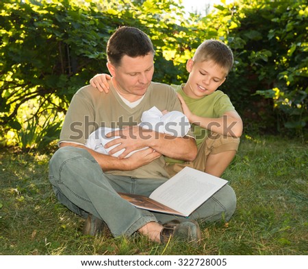 Mature father with a newborn baby in his hands reading book with his elder son on the lawn in the garden. Family reading together. Summer holiday. Happy family concept.