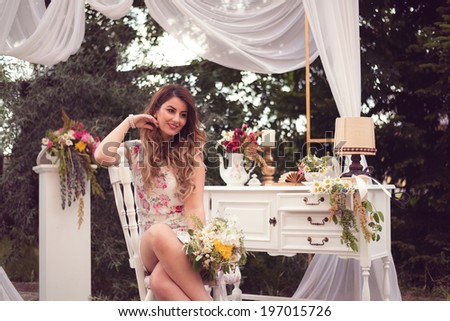 Beautiful woman sitting on a white vintage chair, holding a bouquet, in a shabby chic, vintage decor. White table with vintage objects