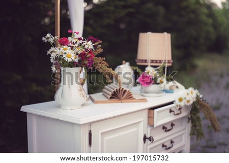Shabby chic decor, white table with vintage objects on it, flowers vase, flower pot an opened book and a lamp, outdoor