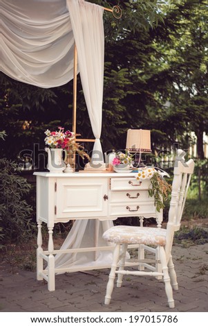 Shabby chic decor, white table with vintage objects on it, flowers vase, flower pot an opened book and a lamp, and a white vintage chair outdoor