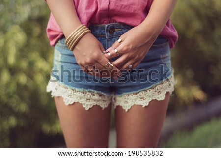 Sexy and attractive woman legs and hands, wearing sexy casual denim shorts with macrame, lace attachment a pink shirt and her hands full of gold fashion accessories rings bracelet