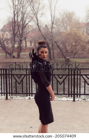 Gorgeous woman wearing a Black Swan feather Collar and a very sexy fashion tight dress with leather sleeves posing at the river side with wrought iron fence in the back