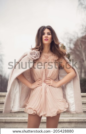 Elegant lady posing on old stairs with her hands in her waist, dressed in a very sexy short nude dress and a warm coat with rose