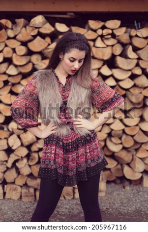 Close up portrait of Fashion beauty posing with attitude in front of a pile of wood wearing a beige fur vest, colorful dress and black tights. Wood background