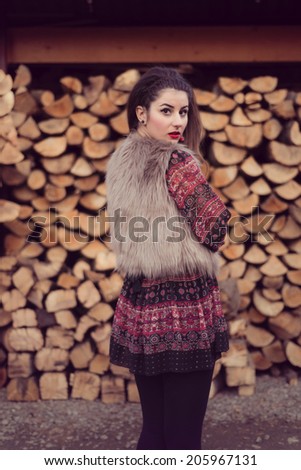 Fashion beauty posing with her back in front of a pile of wood wearing a beige fur vest, colorful dress and black tights. Wood background