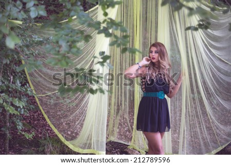 Woman posing into the woods under a green veil canopy, wearing a sexy little black dress