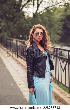 Gorgeous blonde posing in a sexy light blue dress tight and fluid on her body. Fashion shoot of beautiful woman at the river side wearing leather jacket