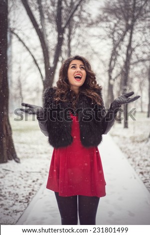 Winter outdoor fashion shoot of attractive blonde in a romantic look, red dress, black fur and sexy tights looking at the falling snow, arms lifted