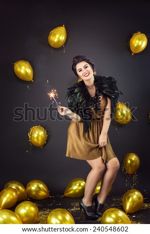 Happy fashion woman dancing, holding fireworks dressed in a gold dress and feathers collar, surrounded with yellow balloons. New Year\'s Eve Party
