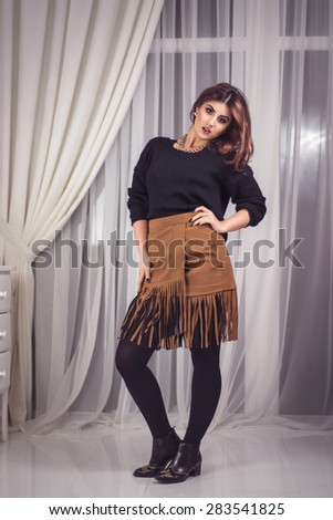 Studio shoot of beautiful fashion woman posing full length dressed with a knitted black sweater and a brown skirt with fringes. Professional make-up