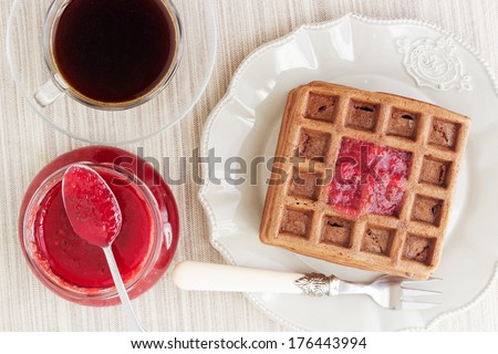A bunch of chocolate gold brown waffles with a bite on light pastel vintage plate and natural napkin. Decorated with red jam, served with a transparent cup of coffee. Photo is taken from the up.